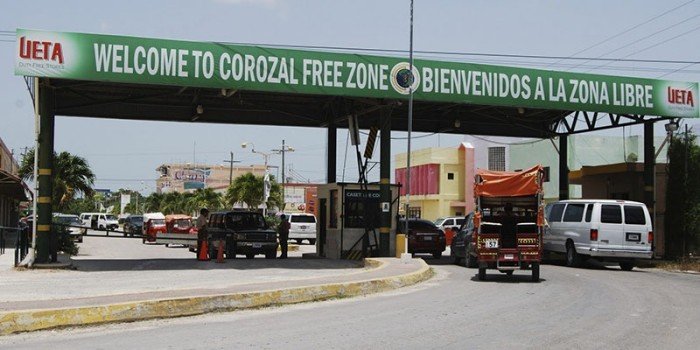 A large green overhead sign reading Welcome to Corozal Free Zone and Bienvenidos a la Zona Libre spans a road where several vehicles are stopped at a checkpoint booth.