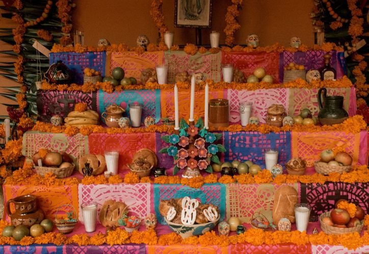 A multi-tiered Day of the Dead altar decorated with orange marigolds, colorful papel picado, candles, fruit, bread, and sugar skulls.