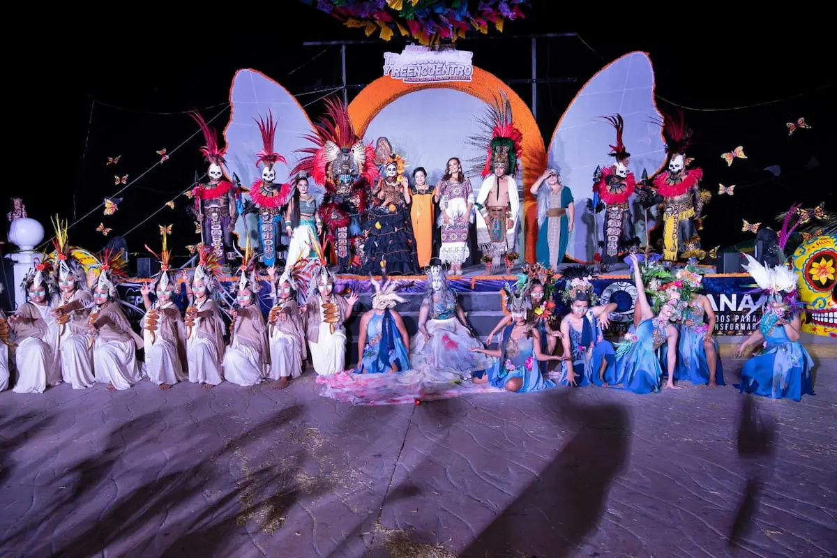 A large group of costumed performers, many wearing skull makeup and feathered headdresses, pose on and in front of an outdoor stage decorated with butterfly cutouts and a sign reading Greencentro.