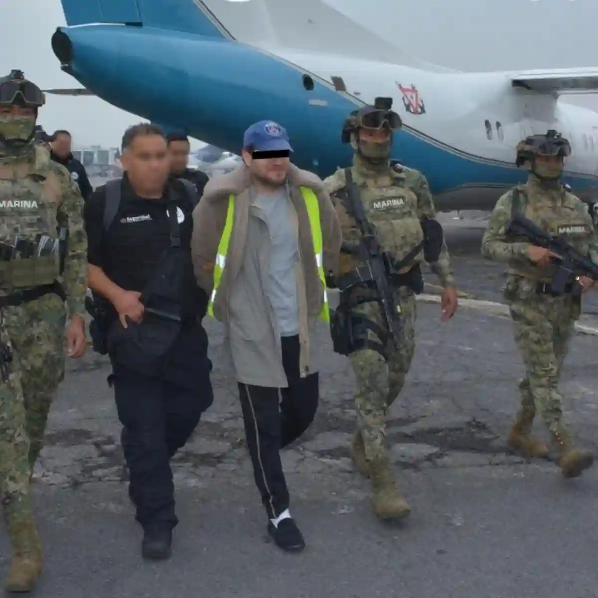 A detained individual is escorted by armed security personnel at an airport.$# CAPTION
