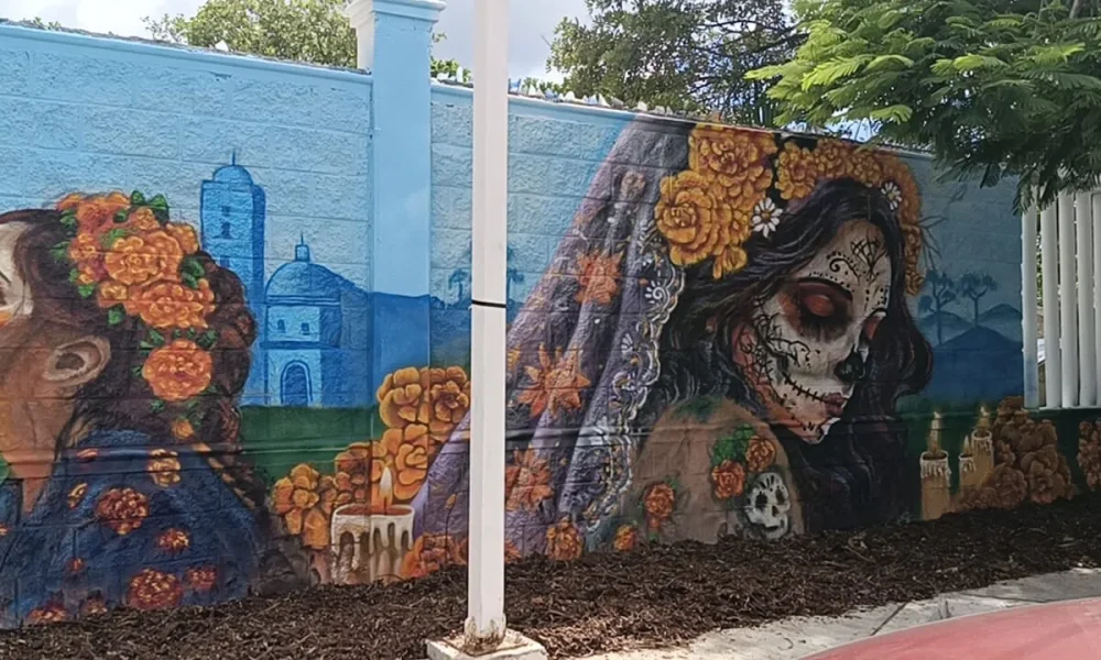 A large outdoor mural painted on a light blue wall depicts two women with orange flower crowns, one with a sugar skull face, alongside a candle, a church dome, and a white pole in the foreground.