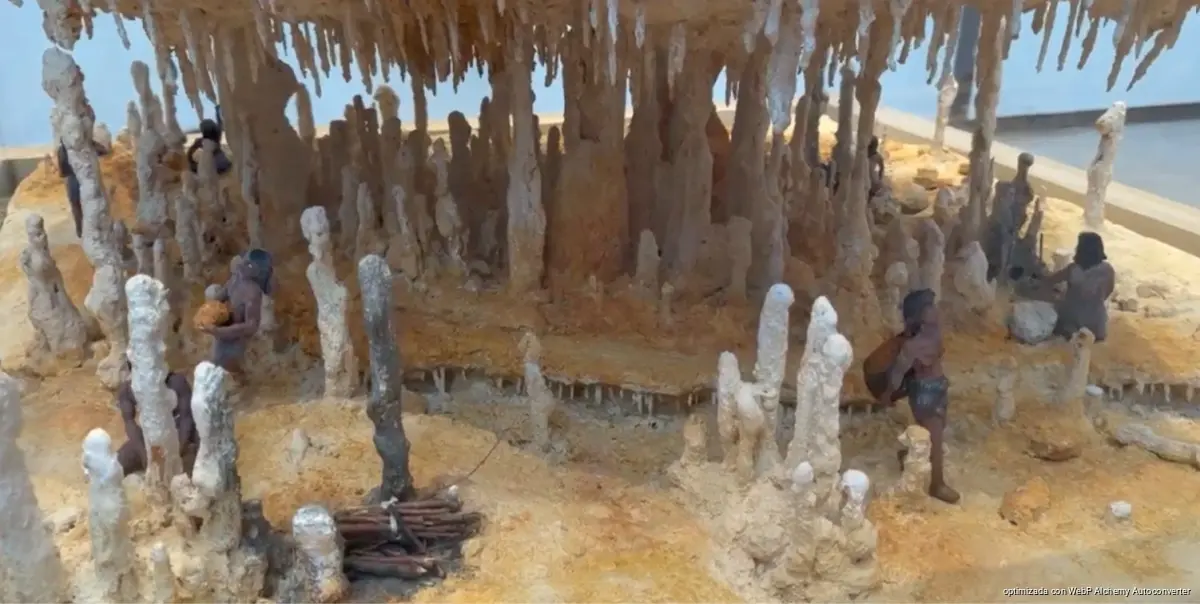 A miniature diorama shows small dark figures standing among pale stalagmites and stalactites on a sandy colored surface.