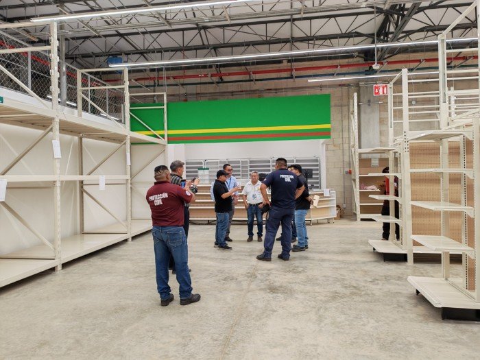 A group of people stand in a large warehouse with empty white metal shelving units on a concrete floor beneath a high ceiling with exposed beams and fluorescent lights.