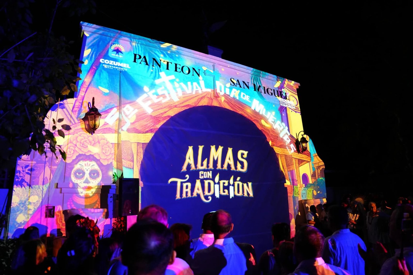A large illuminated projection on a building facade displays the words Almas con Tradicion and Festival de Dia de Muertos above a crowd of people at night.