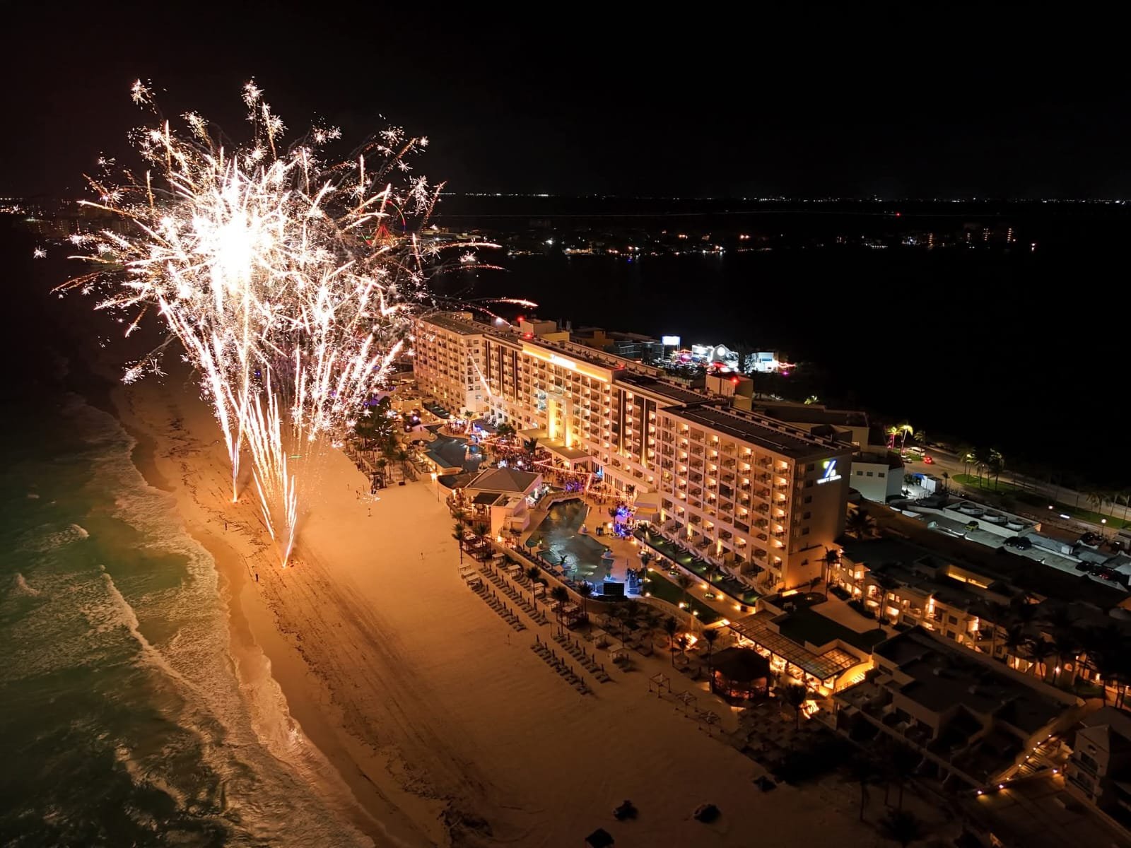 An aerial night view shows a large white fireworks burst above a sandy beach beside a lit multi-story beachfront hotel with pools and rows of lounge chairs, with the ocean on the left and dark water and distant lights in the background.