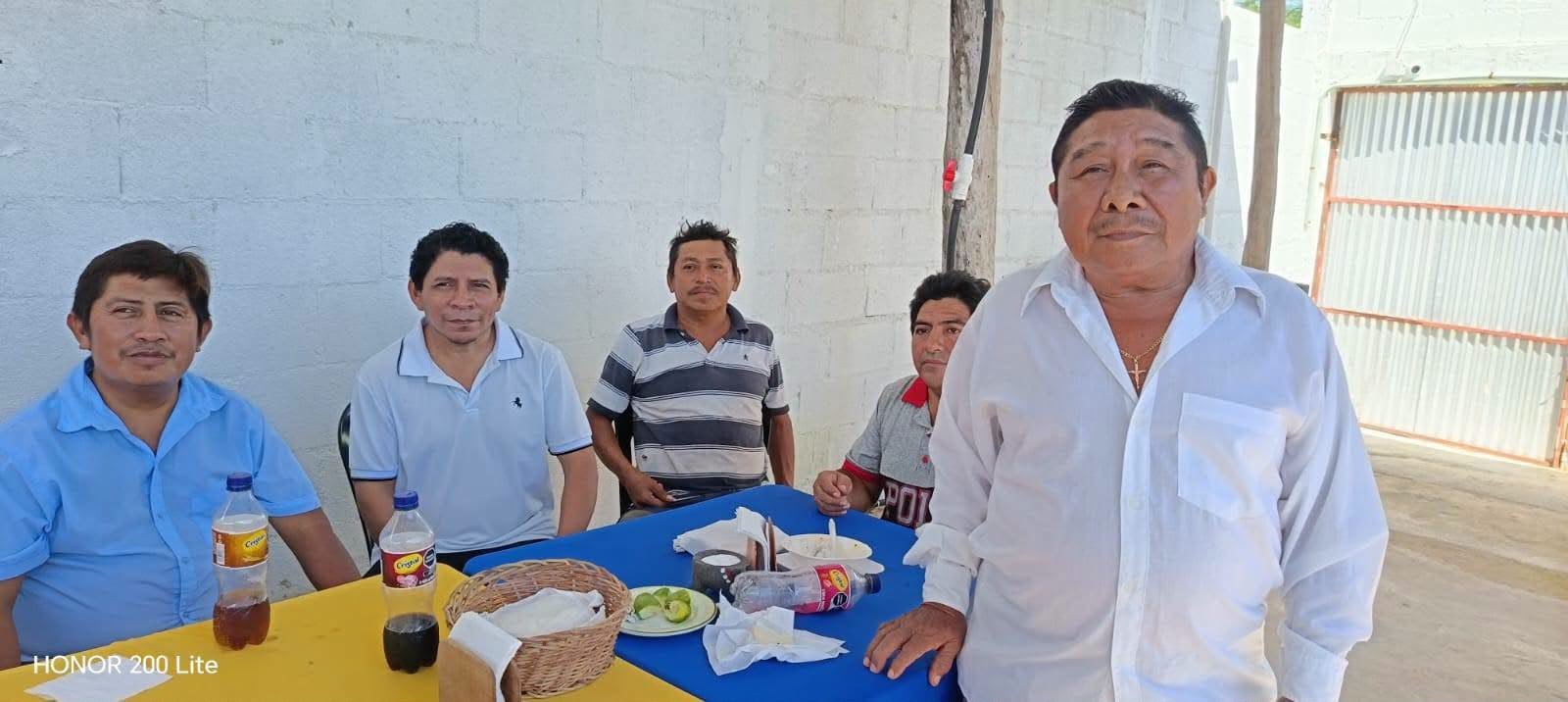 Five men are gathered around a table covered with a yellow and blue cloth, with bottles, a basket, and plates on it, in front of a white block wall.