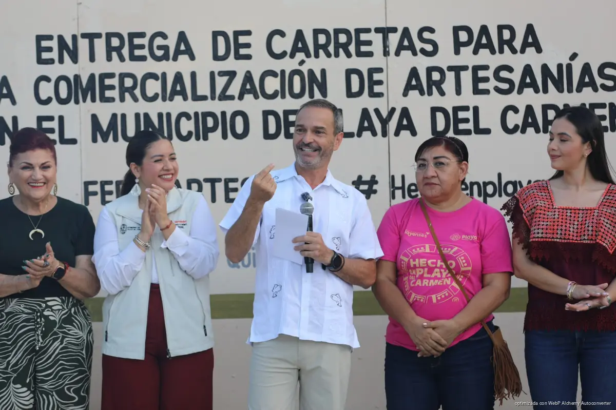 Five people stand in a row in front of a large sign reading Entrega de Carretas para la Comercialización de Artesanías en el Municipio de Playa del Carmen, with a man in a white shirt holding a microphone and paper in the center.