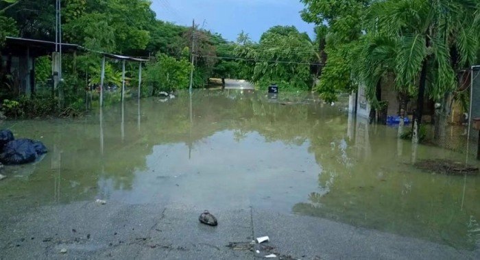 A paved road leads into a large expanse of murky floodwater that surrounds partially submerged structures and trees under a blue sky.