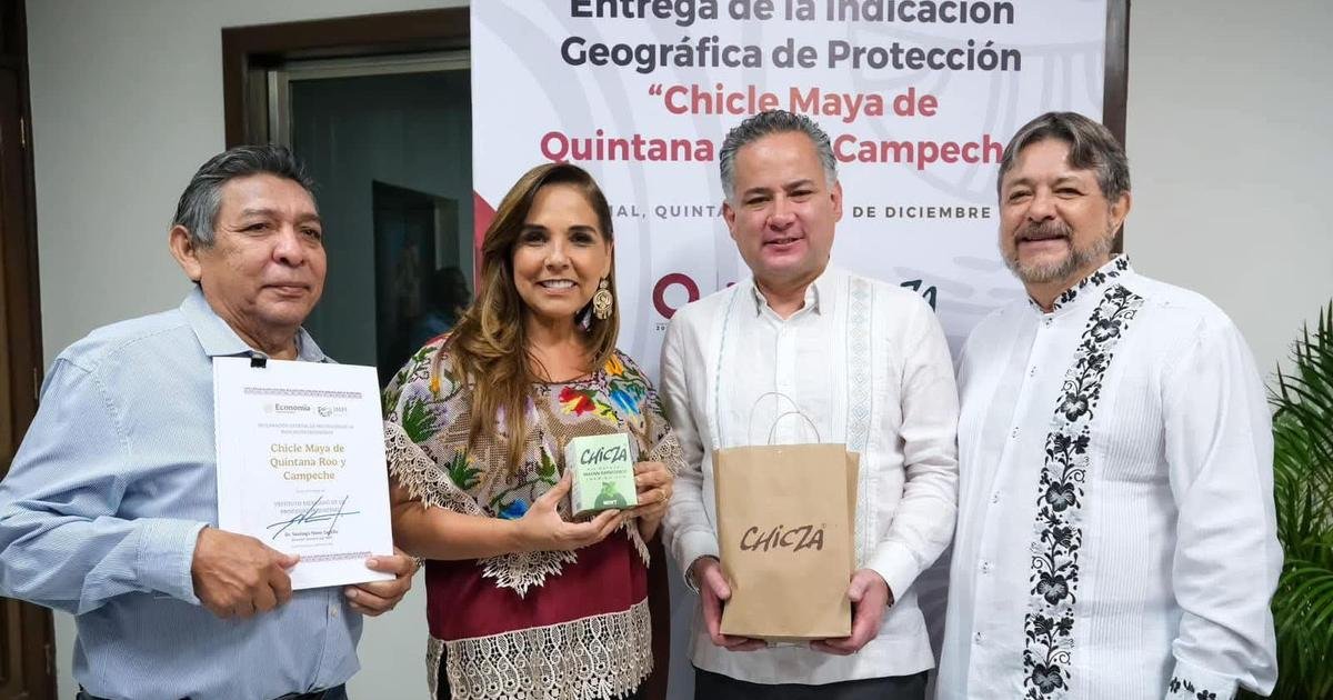 Four people stand in front of a banner reading Entrega de la Indicacion Geografica de Proteccion Chicle Maya de Quintana Roo y Campeche, with one man holding a signed certificate, a woman holding a small green box, and another man holding a brown paper bag labeled CAICZA.