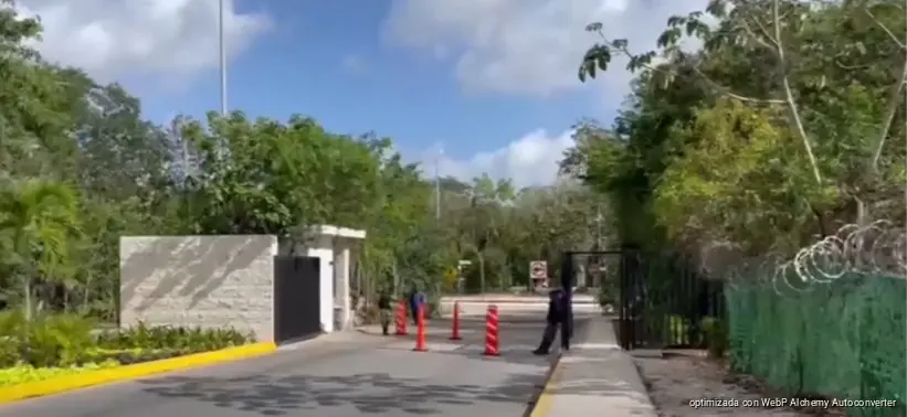 A paved driveway leads to a gated entrance with a stone wall on the left, red and white traffic bollards, a black metal gate, and a person standing near the gate, surrounded by trees under a blue sky.