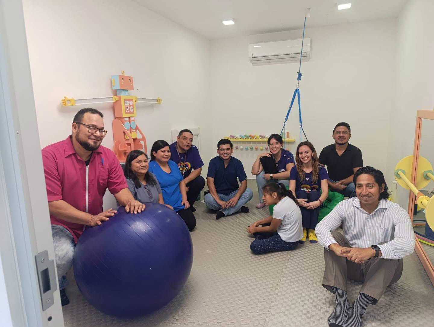 A group of eight adults and one child sit and kneel on the floor of a white room containing a large blue exercise ball, wall-mounted toys, a ceiling hoist and a mirror.