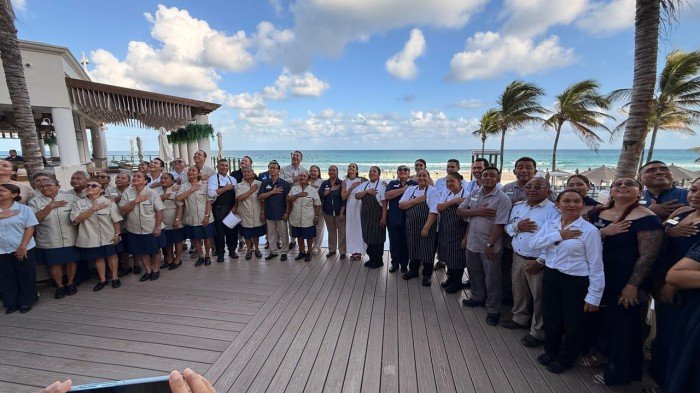 A large group of people in uniforms stand together on a wooden deck with their hands over their hearts, facing a person holding a phone in the foreground, with palm trees and the ocean behind them.