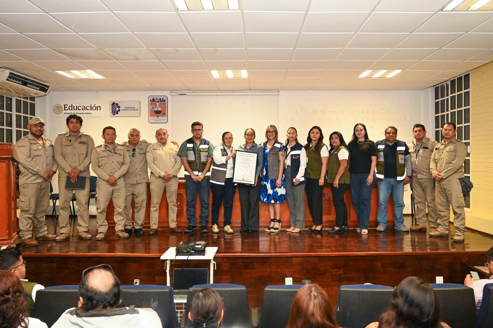 A group of fifteen people in uniforms and civilian clothing stand in a line on a wooden stage, with one person in the center holding a framed certificate, in front of a wall with Education and Tecnologico Nacional de Mexico signs.