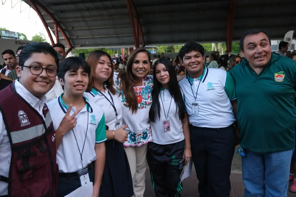 A group of six young people in school uniforms and two adults pose for a photo under a metal roof structure with a crowd behind them.