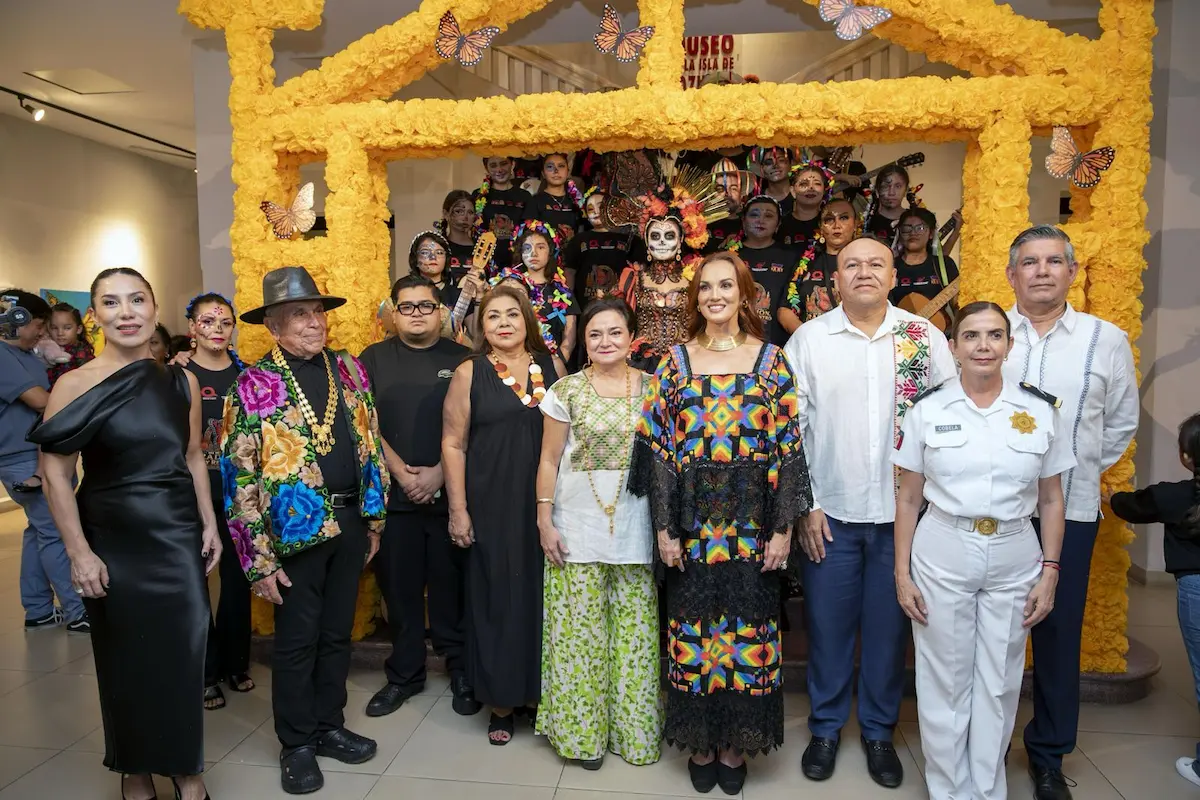A group of adults in formal and traditional dress pose in front of a large yellow floral arch, with children in costume and face paint standing behind them.