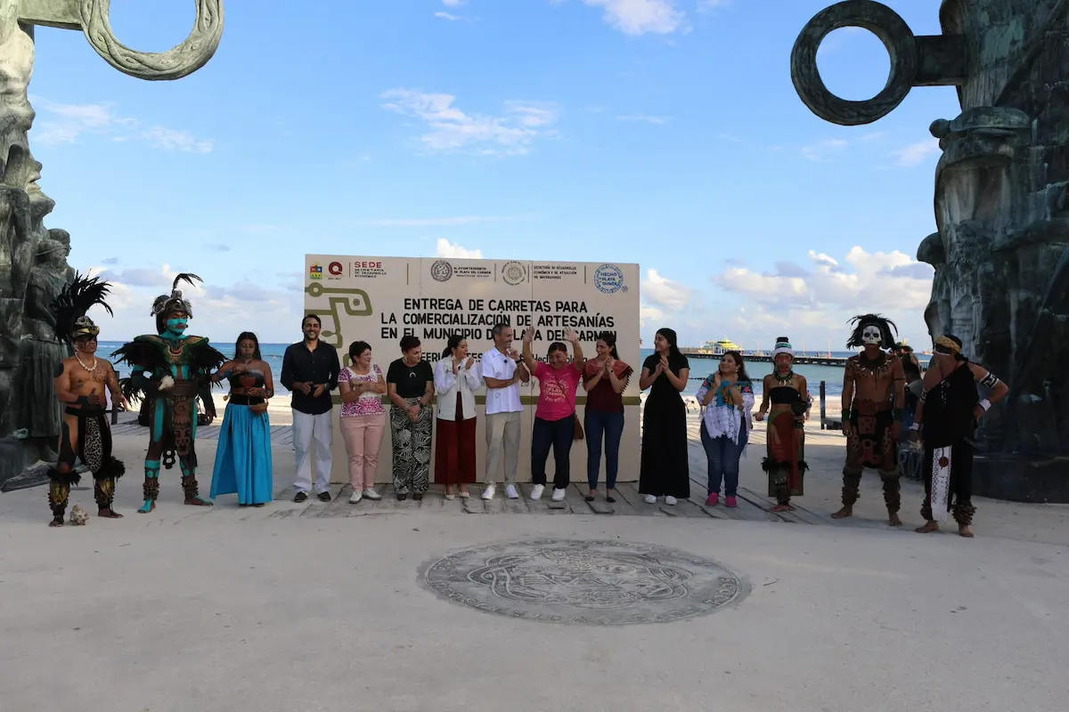 A group of people, some in traditional costumes, stand in a line in front of a sign reading Entrega de Carretas para la Comercializacion de Artesanias en el Municipio de la del Carmen, framed by two large metal sculptures on a sandy plaza.
