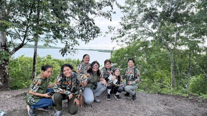 A group of seven women is posing for a photo at a scenic location near a body of water, surrounded by lush greenery.