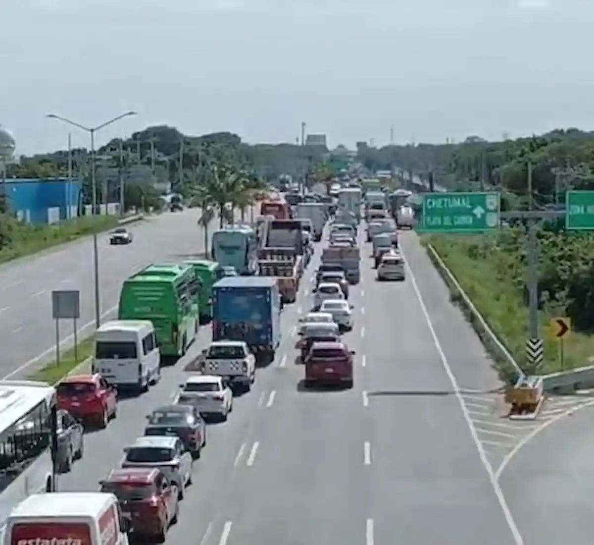 A long line of cars trucks and buses is stopped in heavy traffic on a multi lane highway beside a green overhead road sign reading Chetumal.