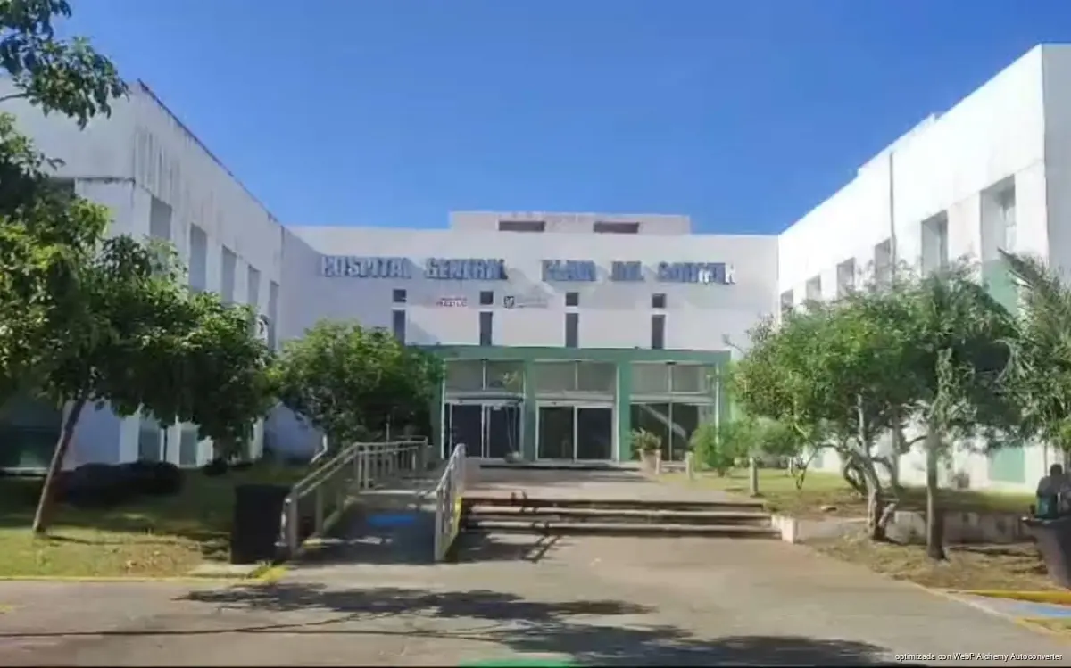 A white multi-story hospital building with the words Hospital General Maria del Carmen on its facade, a green glass entrance, steps, a ramp, and trees in front under a clear blue sky.