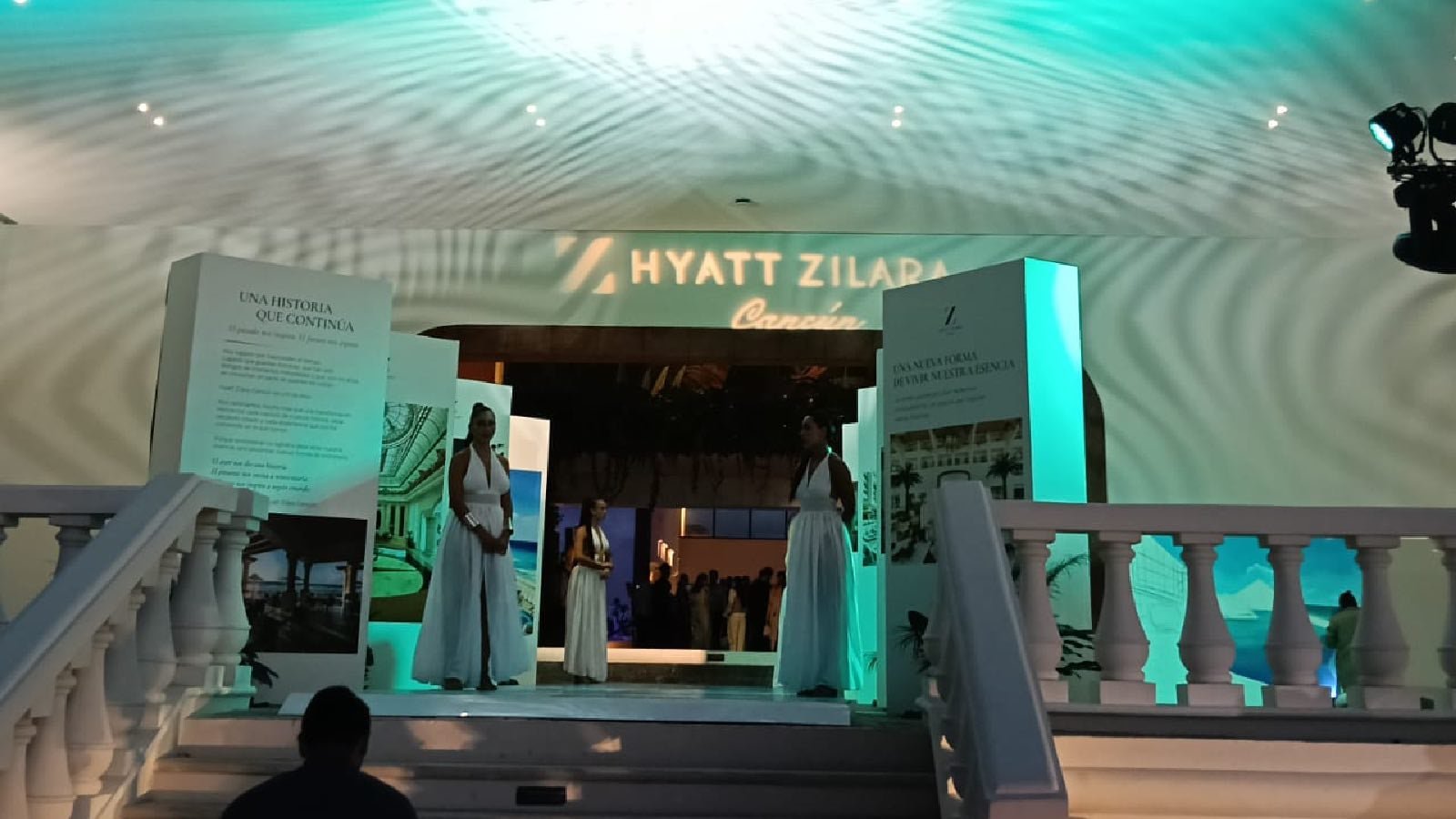 Two women in long white dresses stand on a raised platform between illuminated display panels beneath a Hyatt Zilara Cancun sign, with a white balustrade in the foreground.