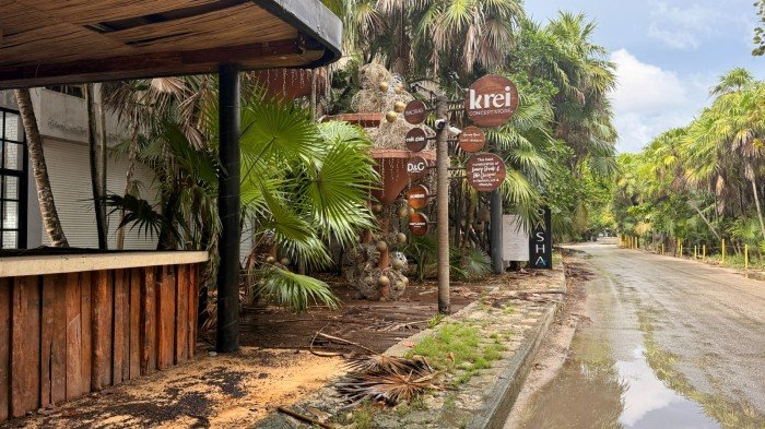 A wooden building with a bar counter stands beside a sign reading krei surrounded by palm trees next to a flooded dirt road.