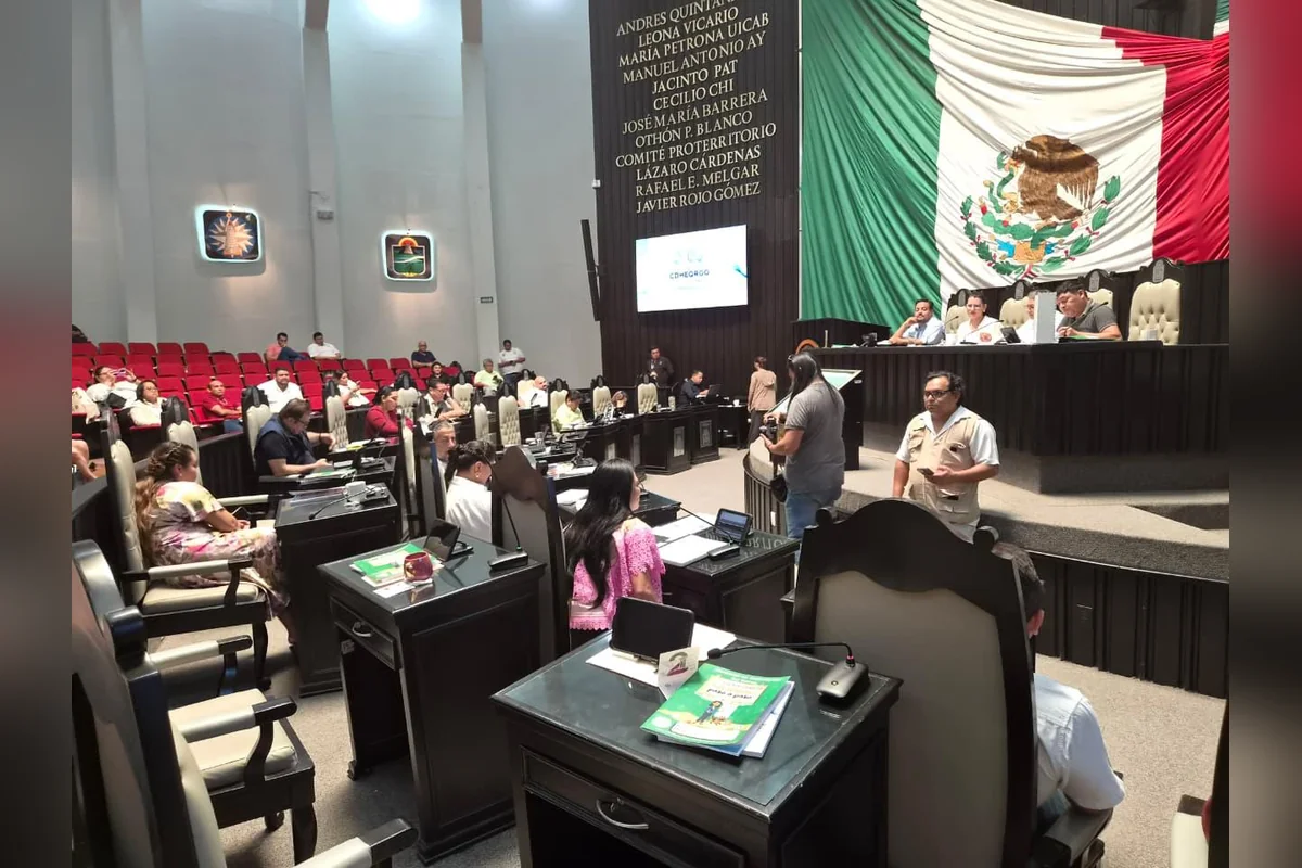 People seated at desks in a large legislative chamber with a Mexican flag and a wall inscribed with names behind the front dais.