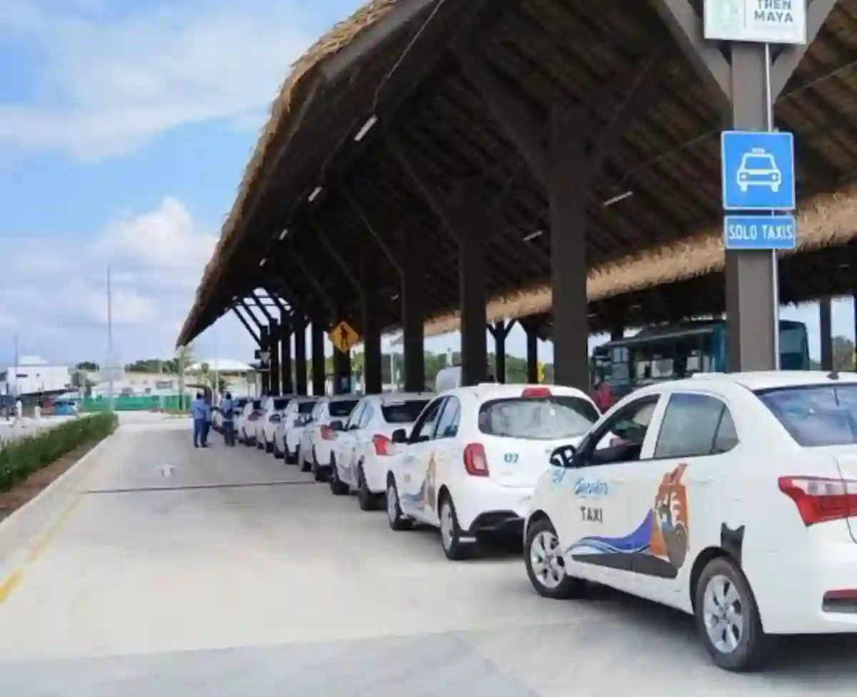 A row of white taxis parked under a structured roof with a sign indicating "Solo Taxis." A couple of people are seen nearby. The sky is partly cloudy.