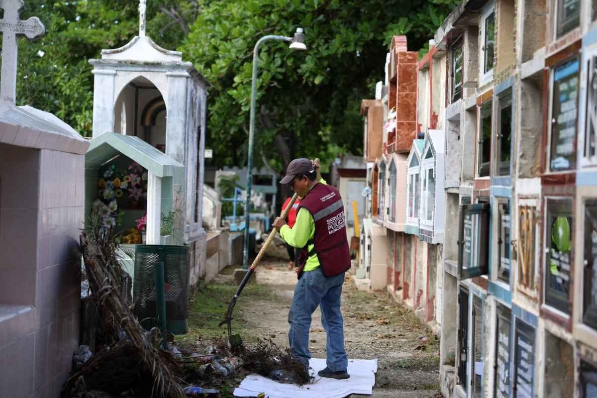 A worker in a cemetery clearing debris with a rake, surrounded by mausoleums and greenery
