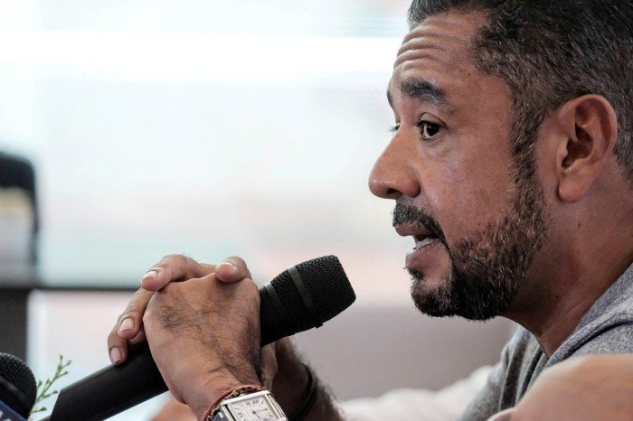 A man with a short beard is shown in profile holding a black microphone with both hands near his mouth.