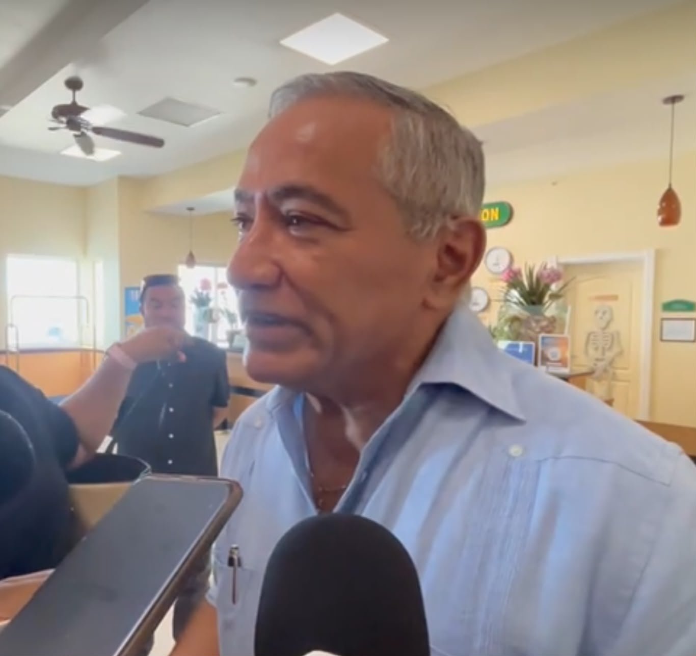 A gray-haired man in a light blue button-down shirt speaks while a microphone and a phone are held toward him in a bright room with a ceiling fan and a skeleton poster in the background.