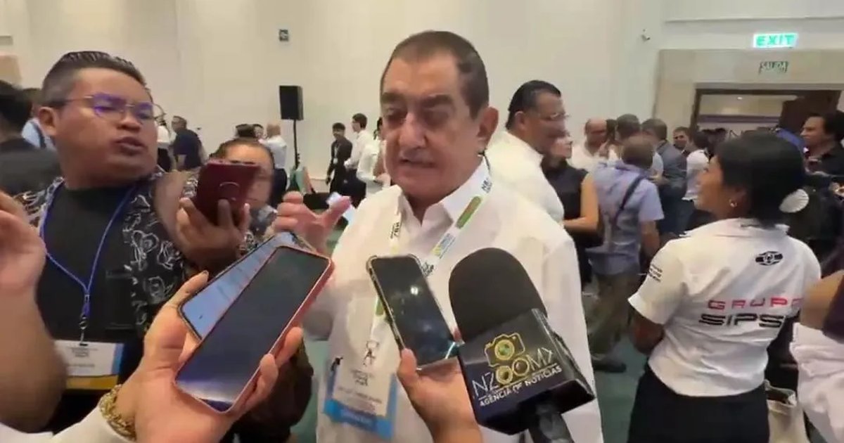 A man in a white shirt and lanyard speaks while several people hold smartphones and a microphone with a Zoom news agency logo toward him in a crowded hall.