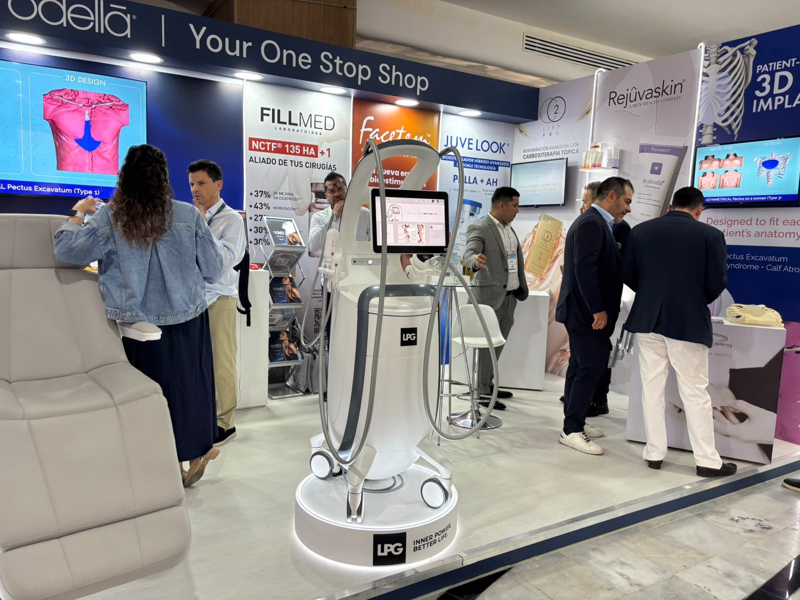 People stand around a white LPG treatment device on a lit circular platform in front of a trade show booth with screens and brand signs including Fillmed, Juvelook and Rejuvaskin.