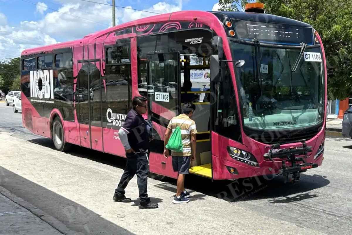 A pink public transport bus with the name "Mobi" on the side is stopped at a bus stop. A passenger is approaching the bus, while another person stands nearby. The scene is set on a sunny day with blue skies and visible trees in the background.$# CAPTION
