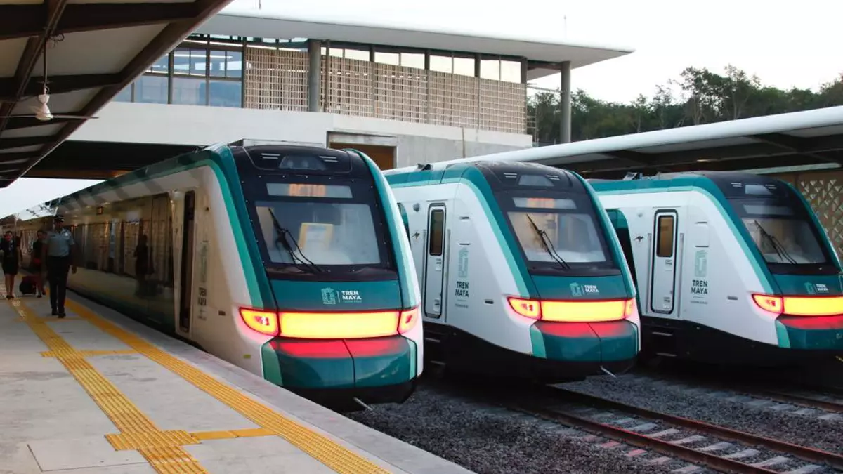 Three modern Tren Maya trains at a station with passengers nearby and a large building above.$# CAPTION