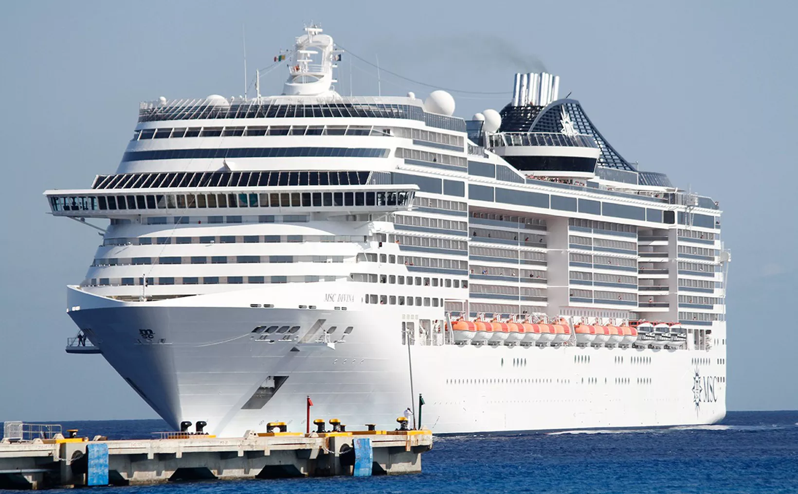 A large white MSC cruise ship with many decks of windows and orange lifeboats is docked beside a concrete pier on blue water.
