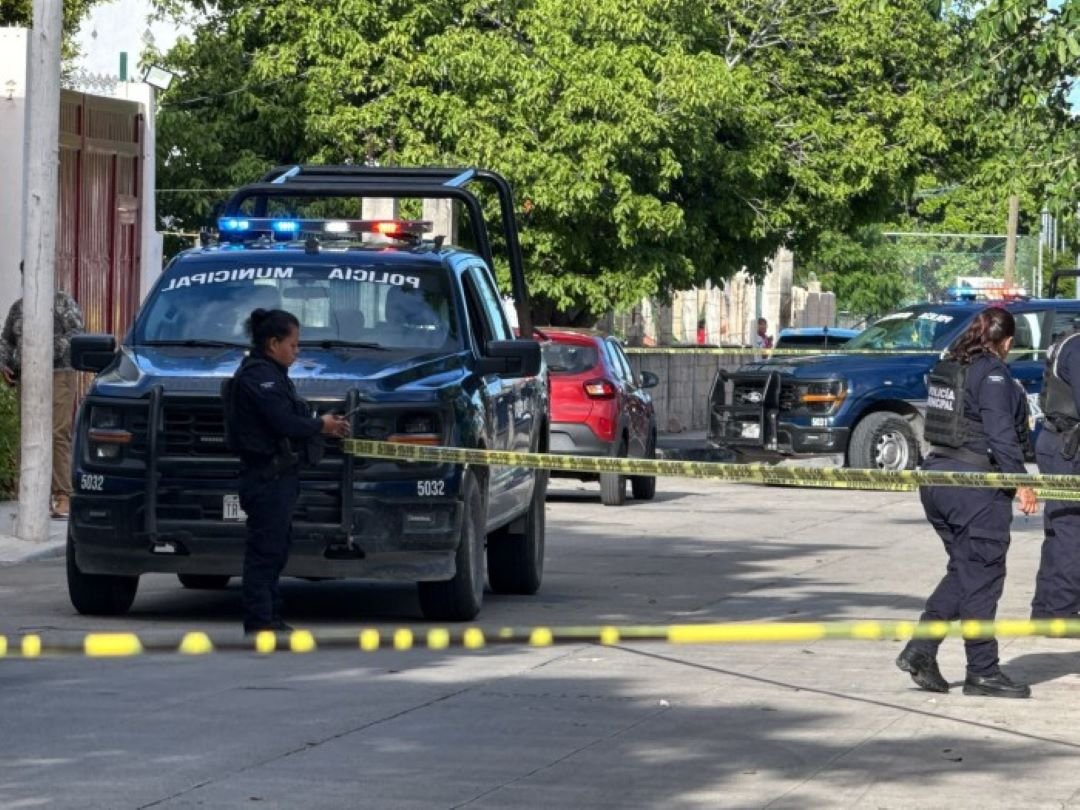 A dark blue municipal police pickup truck with lights on its roof is parked on a street behind yellow caution tape, with officers in dark uniforms standing nearby and a red car and another police truck in the background.
