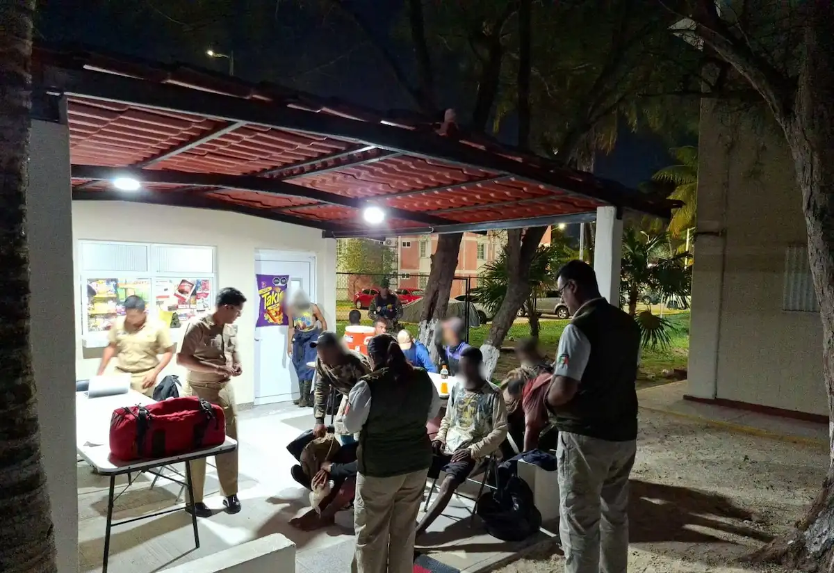 At night under a red corrugated roof, several people in tan uniforms and vests stand and sit around a table holding a large red duffel bag, with a white building and trees behind them.