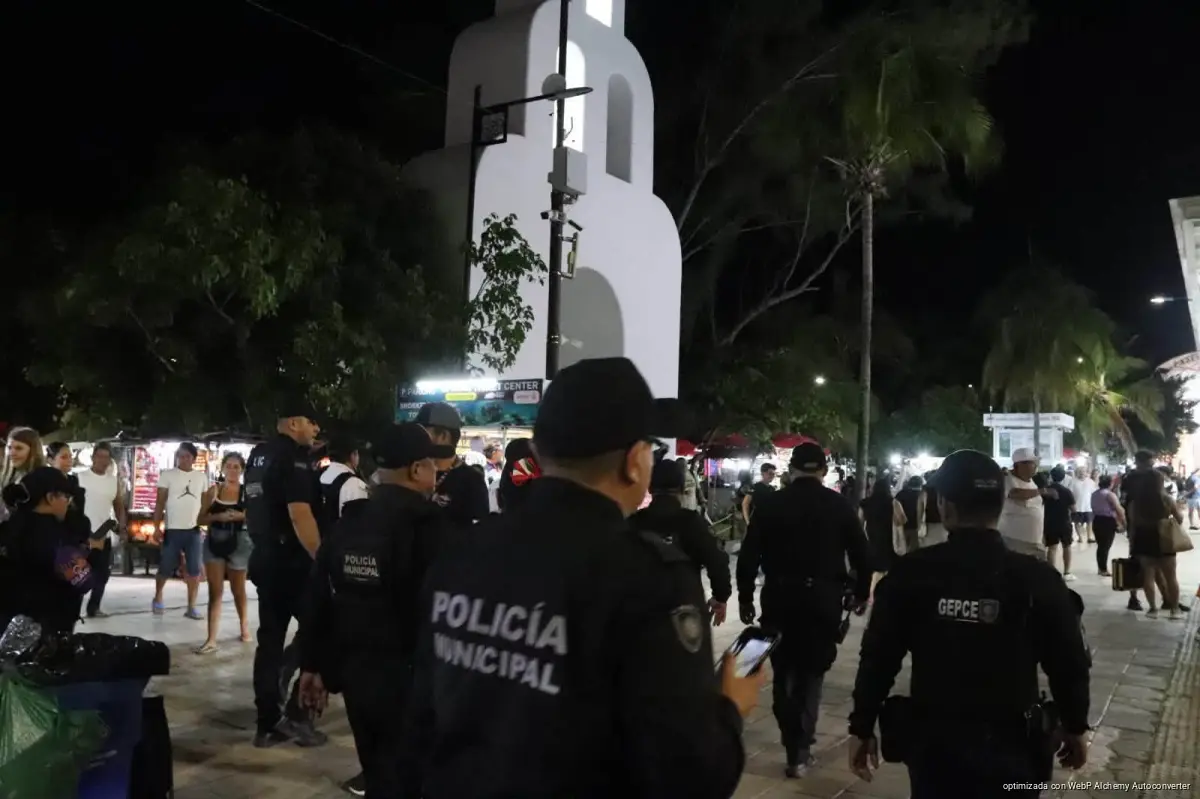 Police officers in dark uniforms with Policia Municipal and GEPCE markings stand among pedestrians at night near a tall white church tower and trees.