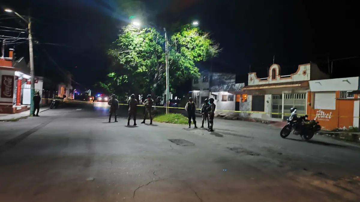 A nighttime view of a paved street where several uniformed officers stand behind yellow police tape near a large tree, with a motorcycle parked on the right and buildings on both sides.