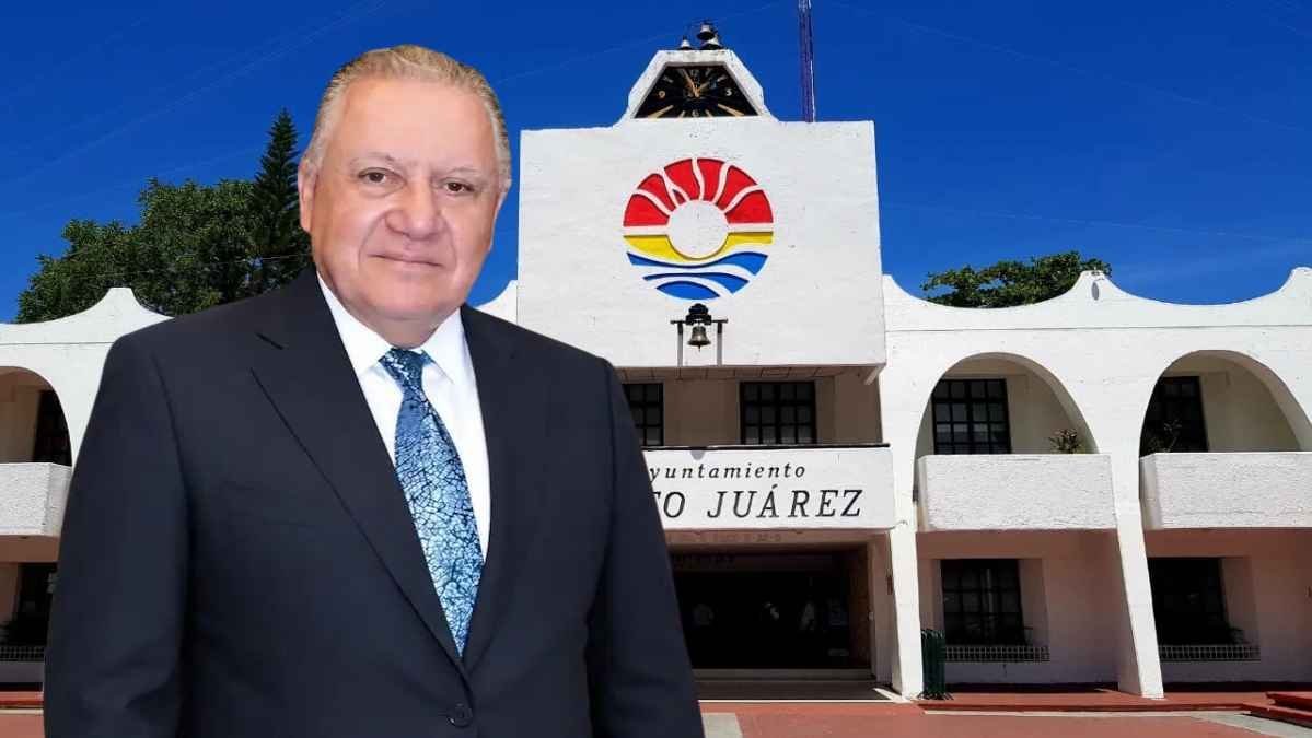 A man in a suit stands in front of the City Hall of Puerto Juárez, featuring a colorful logo and bright blue sky in the background.$#$ CAPTION