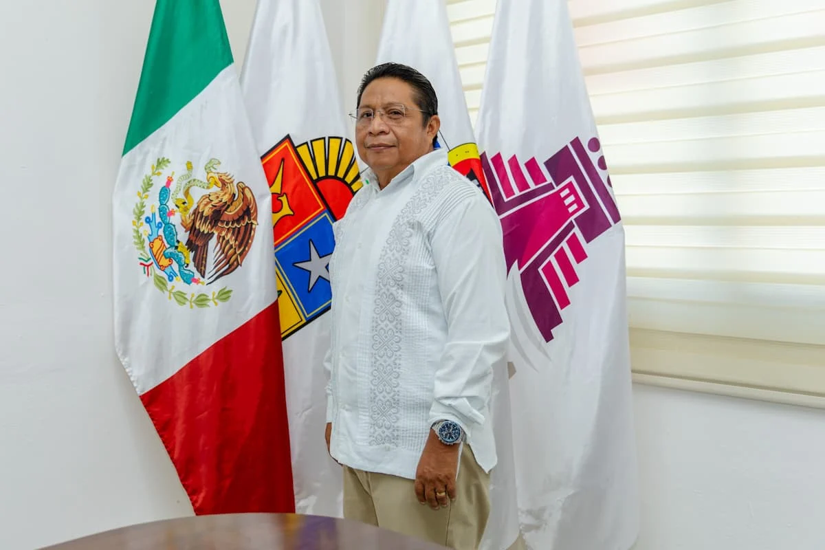 A person in a white shirt stands between three flags, including the Mexican flag, in an official setting