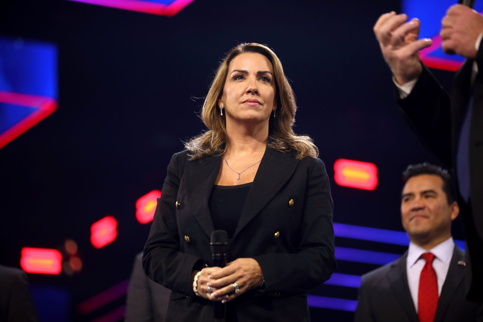 A woman in a black blazer stands confidently on stage, holding a microphone, while a man in formal attire smiles in the background under bright lights.$#$ CAPTION