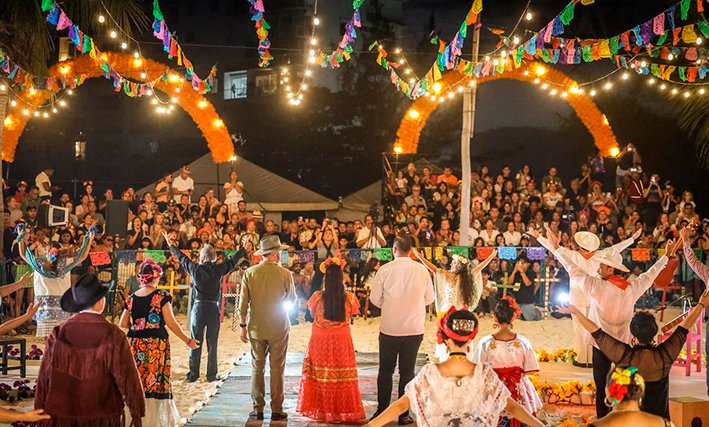 A group of people in traditional Mexican clothing dance in a line on a paved area at night, surrounded by a large seated crowd, with colorful paper banners and string lights overhead.