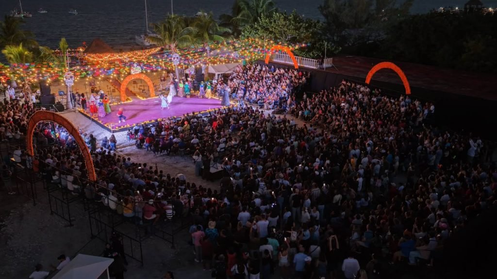 A large crowd watches performers in white costumes on a purple lit stage decorated with orange arches and string lights beside the water at night.