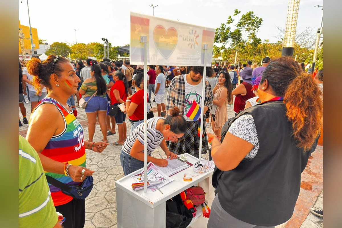 A woman in a rainbow tank top stands beside a small white booth where another woman writes on a clipboard, with a crowd of people and a rainbow heart banner behind them.