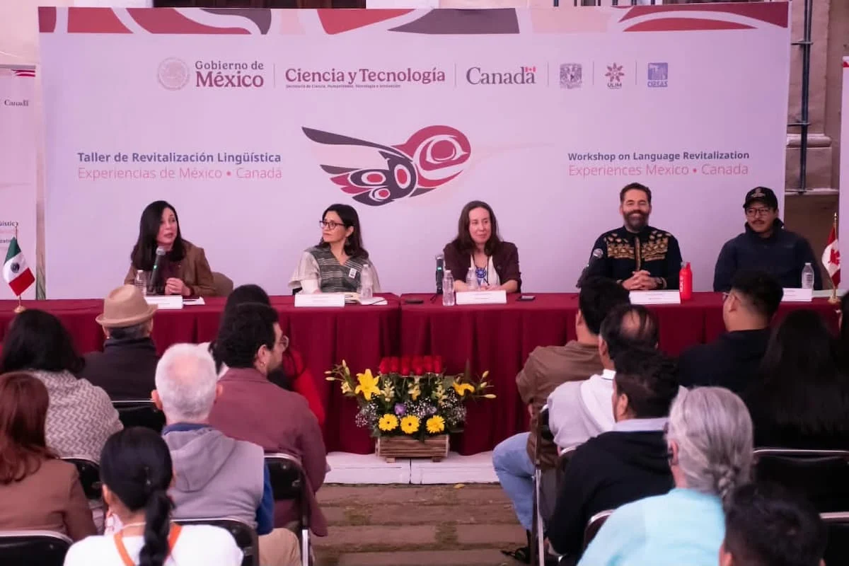 Five panelists sit behind a red draped table on a stage in front of a large banner reading Taller de Revitalización Lingüística and Workshop on Language Revitalization, with an audience seated in the foreground.