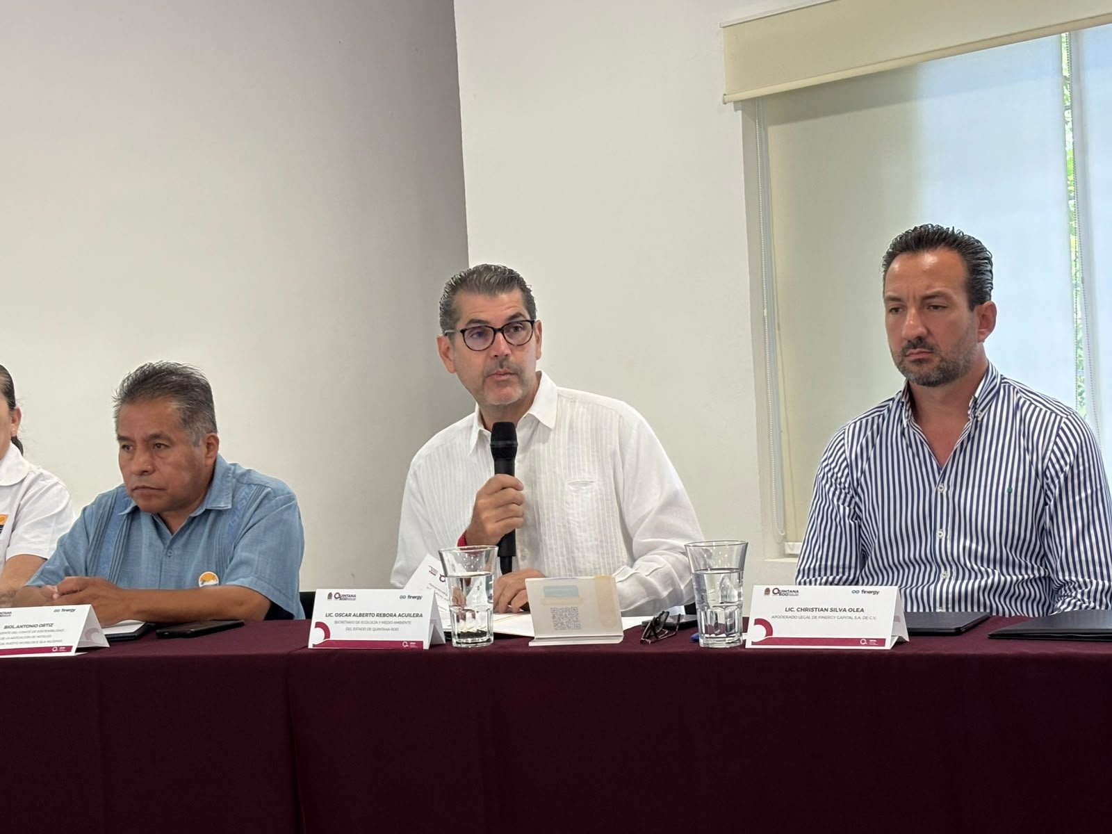 Three men sit behind a maroon-draped table with name cards and water glasses, and the man in the center holds a black microphone.