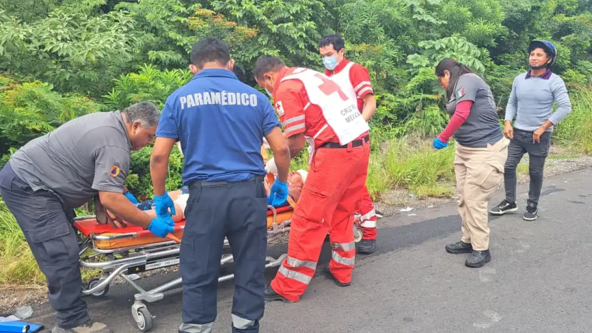 Several paramedics in blue and red uniforms, one wearing a vest marked Cruz Roja Mexicana, stand around a wheeled stretcher holding a patient on a paved road beside dense green vegetation, while a person in a helmet and a woman in a grey shirt look on.