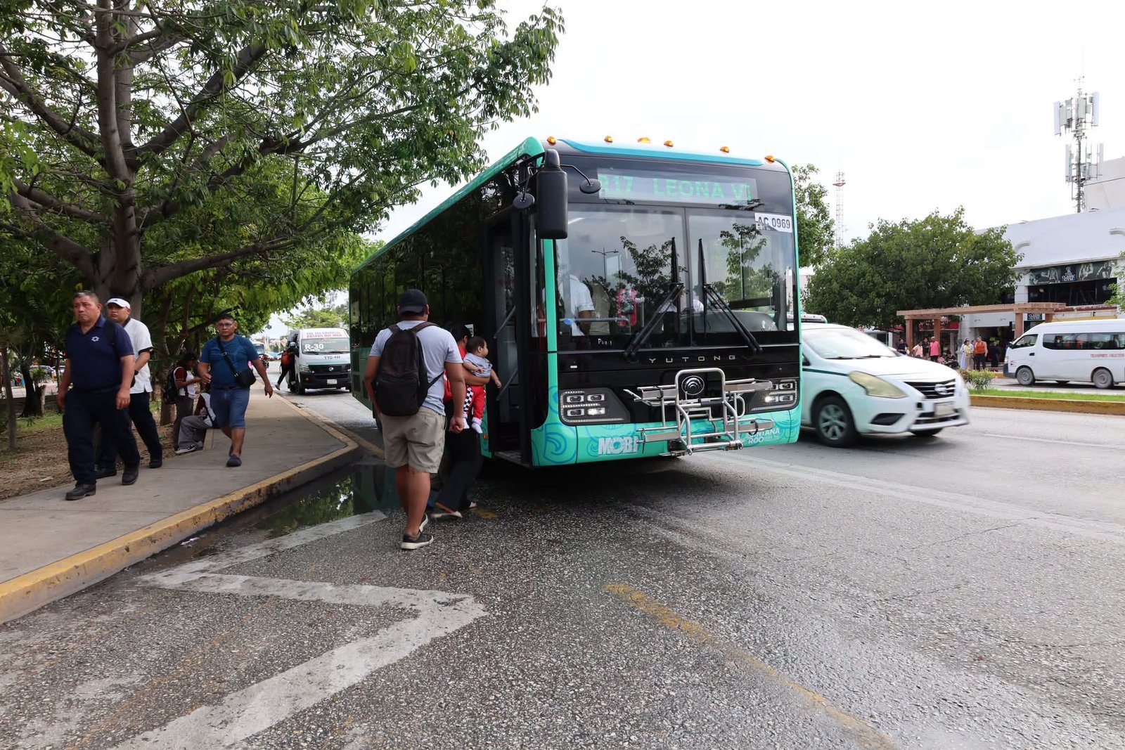People walk on a sidewalk and approach the open door of a turquoise Yutong bus stopped beside a wet curb, with a white car and other vehicles on the road behind it.