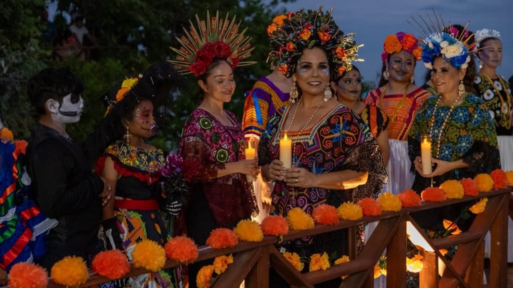 A group of people wearing colorful embroidered traditional clothing and floral headdresses stand behind a wooden railing decorated with orange and yellow flowers, several holding lit candles, with one person at left wearing skull face paint.